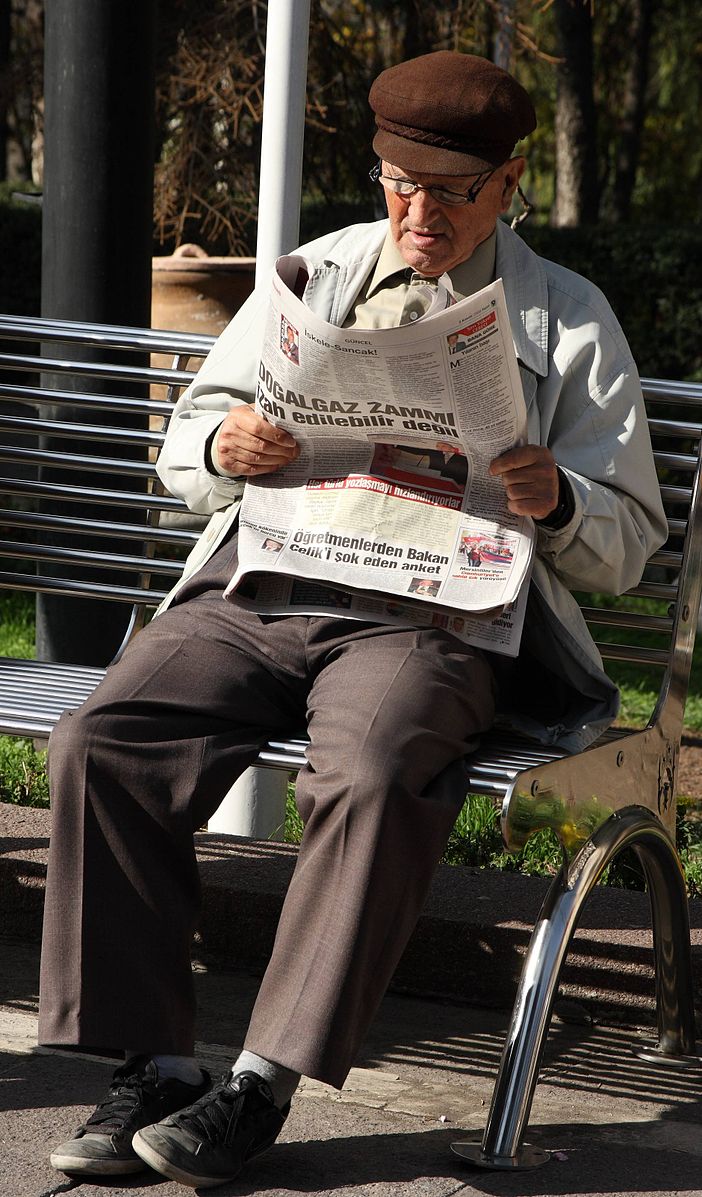 This picture shows a man reading a newspaper while seated on a park bench.
