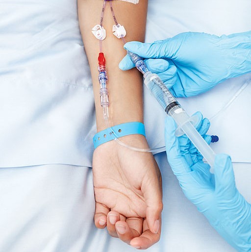 This picture shows the arm of a patient fitted with an IV drip while the gloved hands of a health professional inject clear fluid directly into the bloodstream.