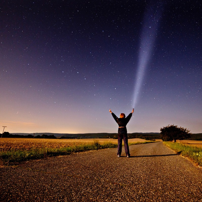 This picture shows a woman standing in the middle of a country road at night and reaching toward the star-filled sky above.