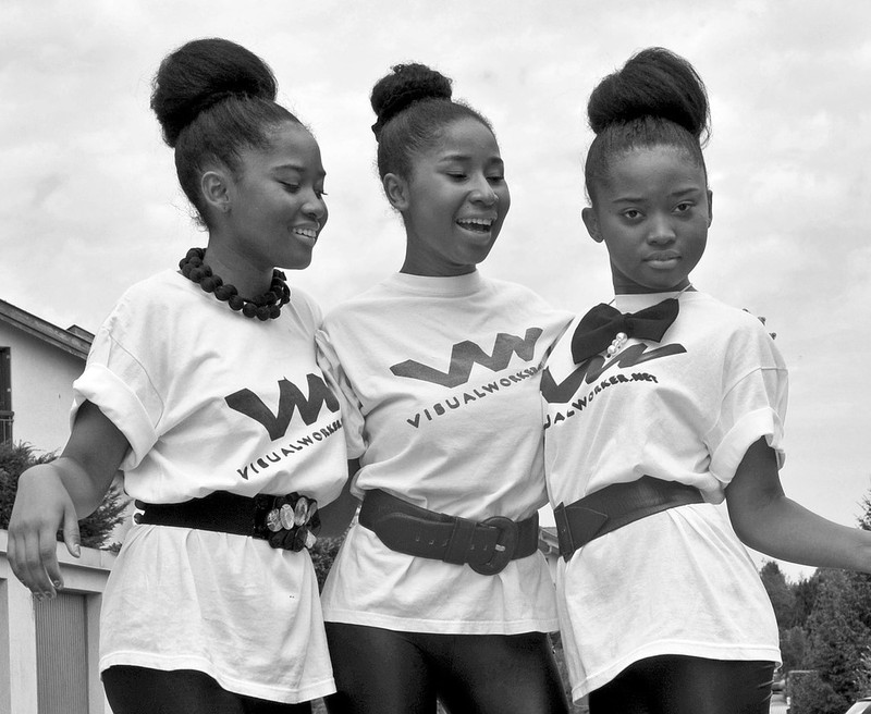This picture shows a group of teen girls with matching shirts and hairstyles posing together with their arms around one another.