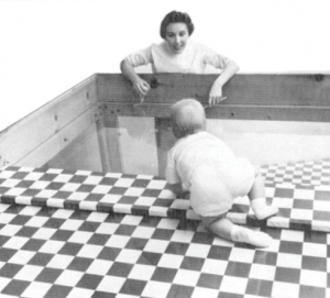 This picture shows a baby crawling to a woman over a surface that is part checkered table cloth and part clear glass.